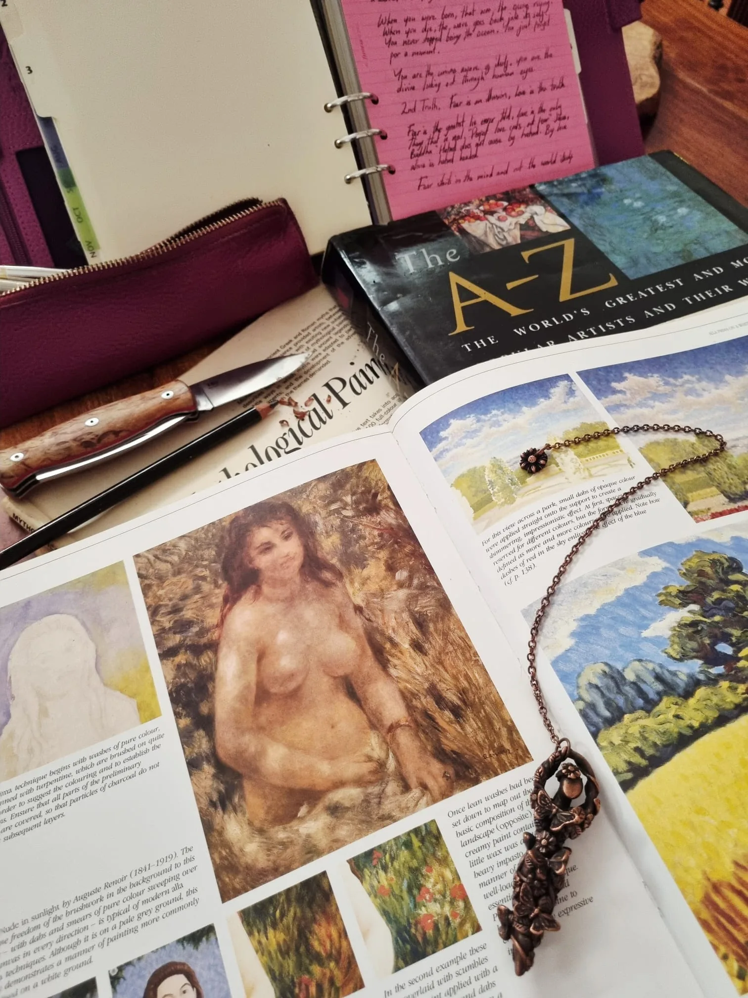 An artist's handwritten notebook and sketchbook beside reference art books and a pendant