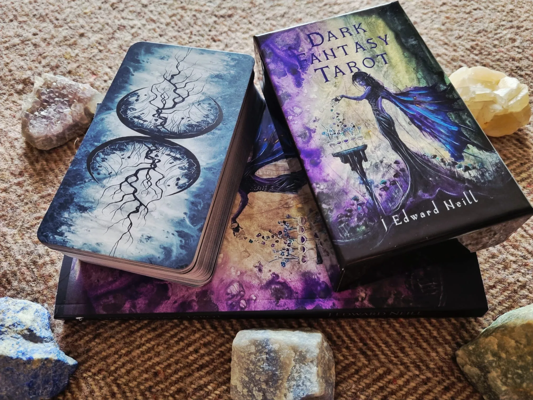 The Dark Fantasy Tarot box, guidebook and deck resting on woven fabric beside amethyst, lapis lazuli and citrine, with the twin moon and root card backs facing up.