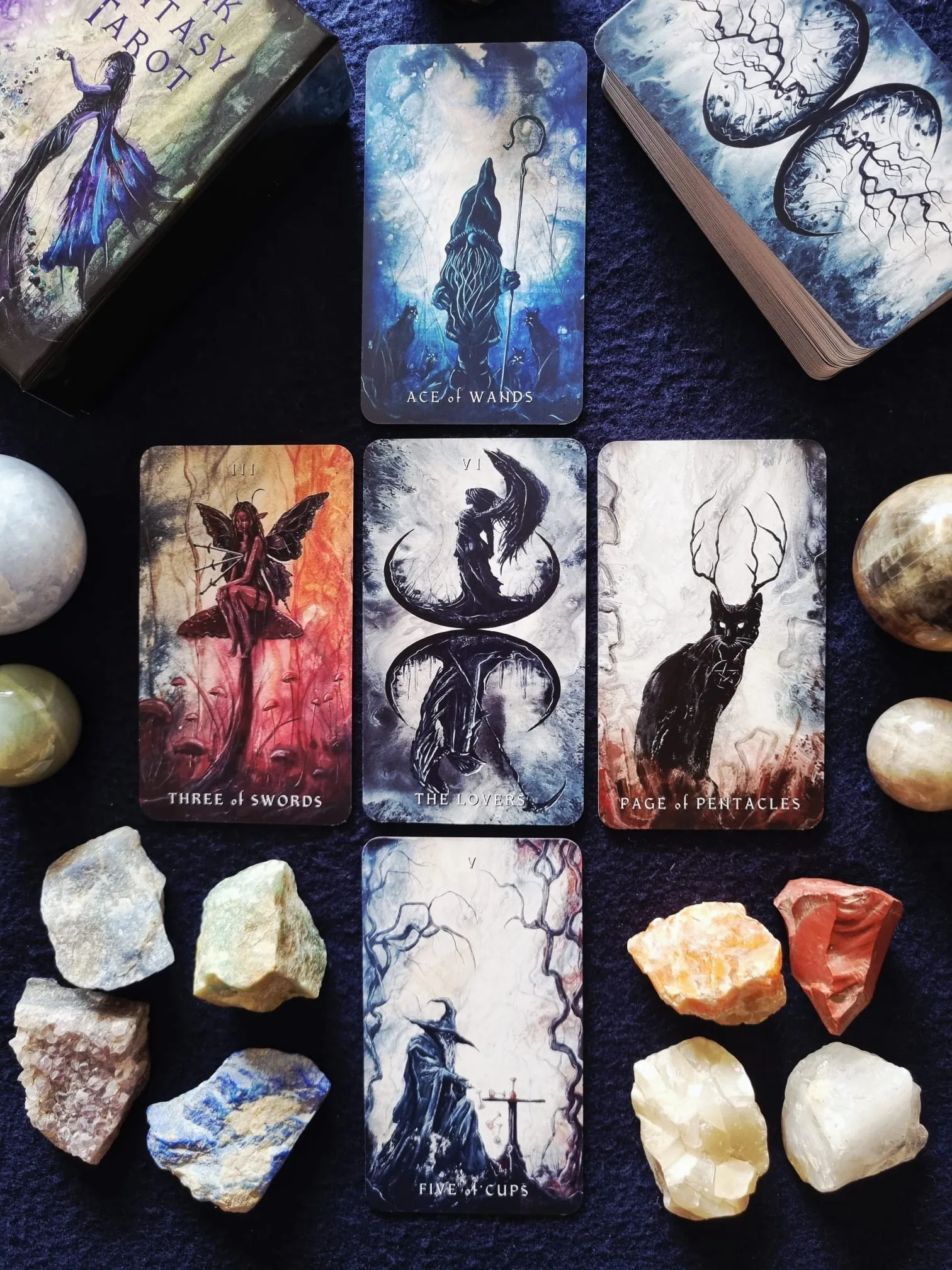 Dark Fantasy Tarot cards arranged in a cross on dark blue velvet, showing the Ace of Wands, Three of Swords, The Lovers, Page of Pentacles and Five of Cups beside crystal spheres.