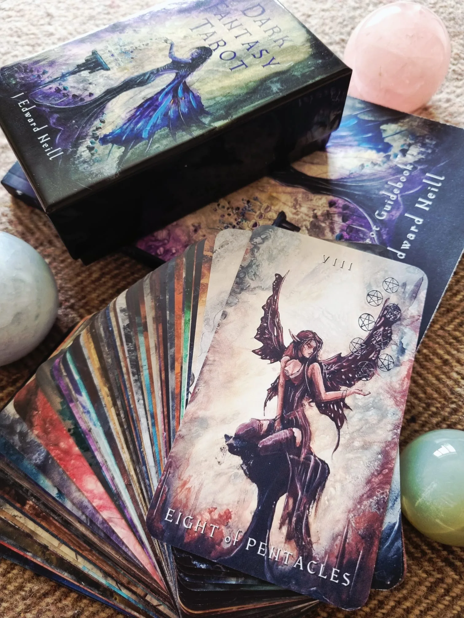 The Eight of Pentacles from the Dark Fantasy Tarot showing a winged faerie in shadow, fanned above the rest of the deck beside the box and guidebook.
