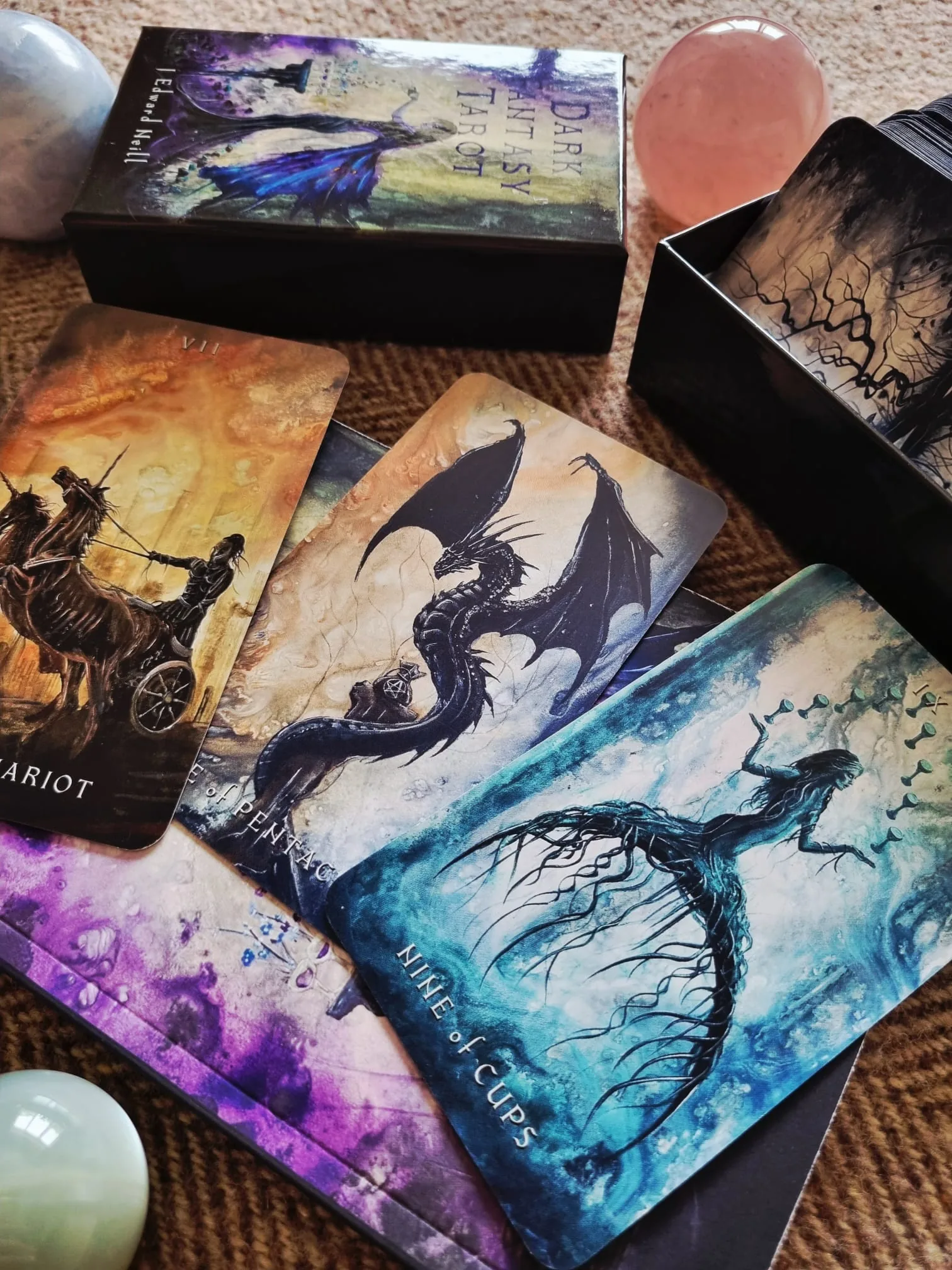 A winged black dragon guarding a pentacle on a Dark Fantasy Tarot card, laid beside The Chariot and the Nine of Cups with the deck box behind.