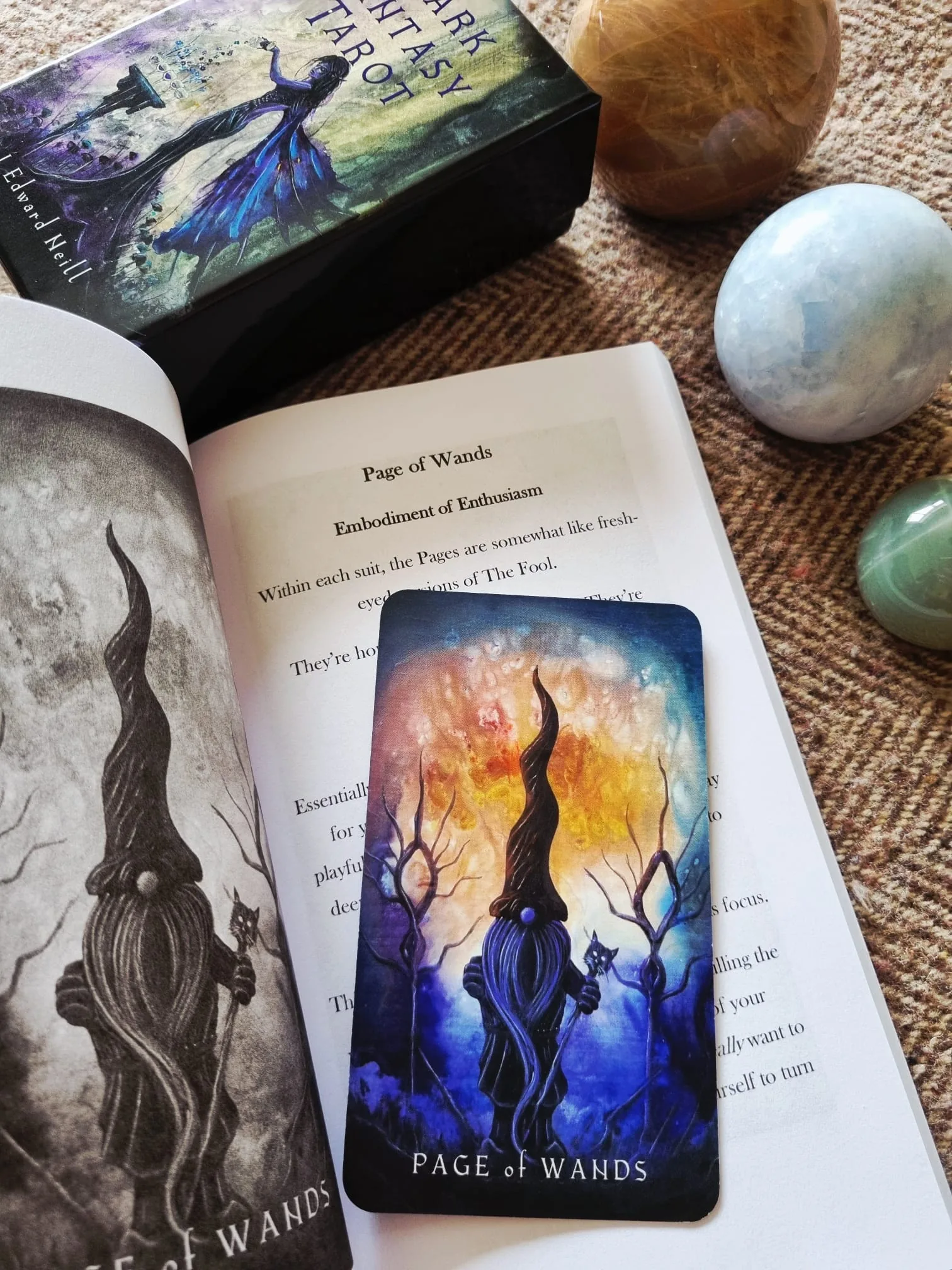 The Dark Fantasy Tarot guidebook open at the Page of Wands entry with the matching card resting on the page, beside the deck box and crystal spheres.