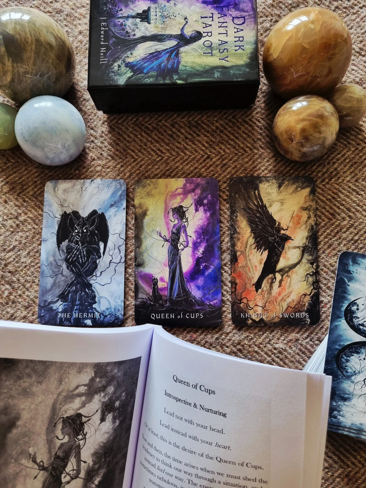 A three card Dark Fantasy Tarot spread of The Hermit, Queen of Cups and Knight of Swords, with the guidebook open at the Queen of Cups page below.