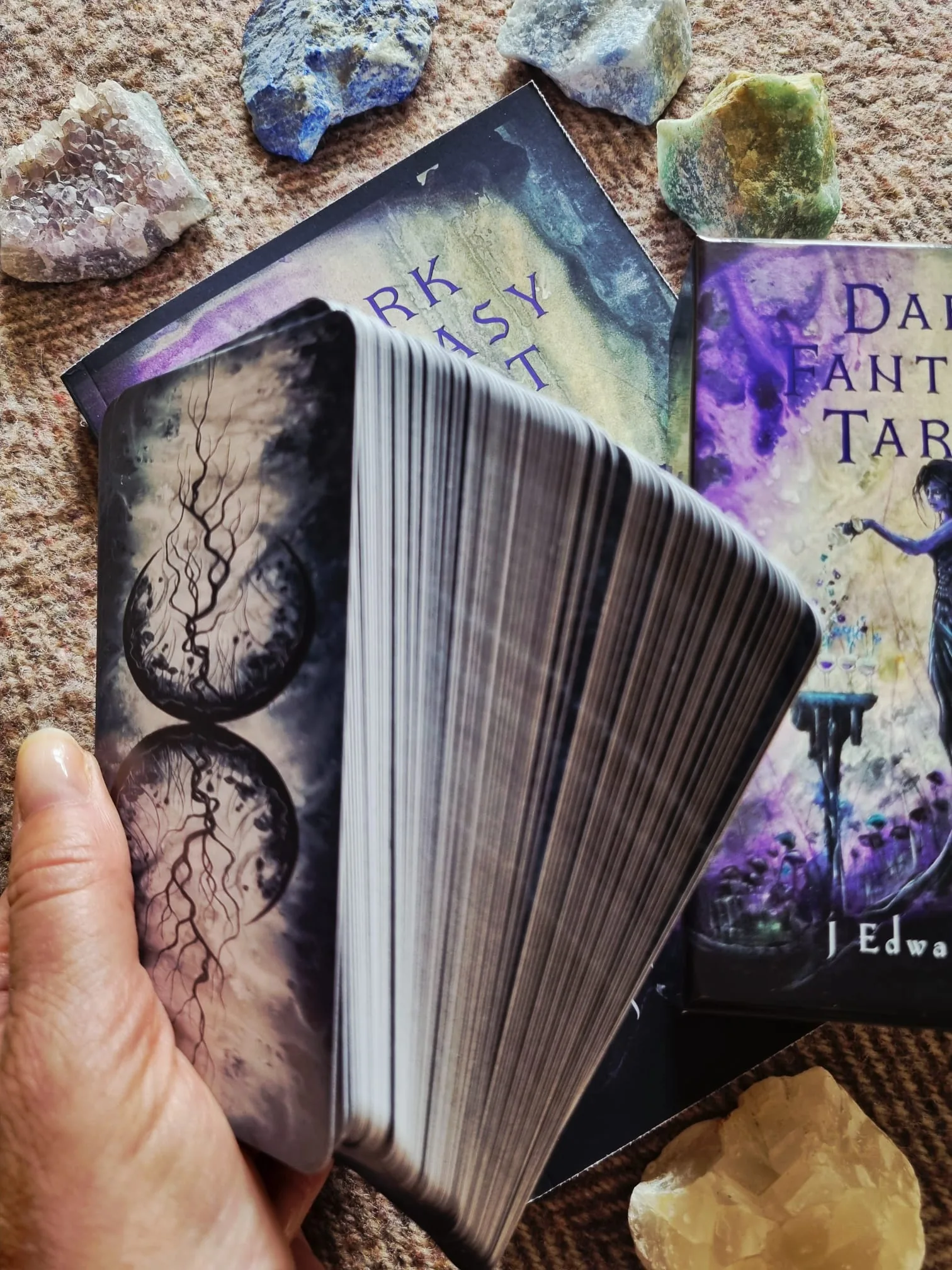 A hand fanning the Dark Fantasy Tarot deck to show the twin moon and root card backs, with the box and guidebook resting on woven fabric behind.
