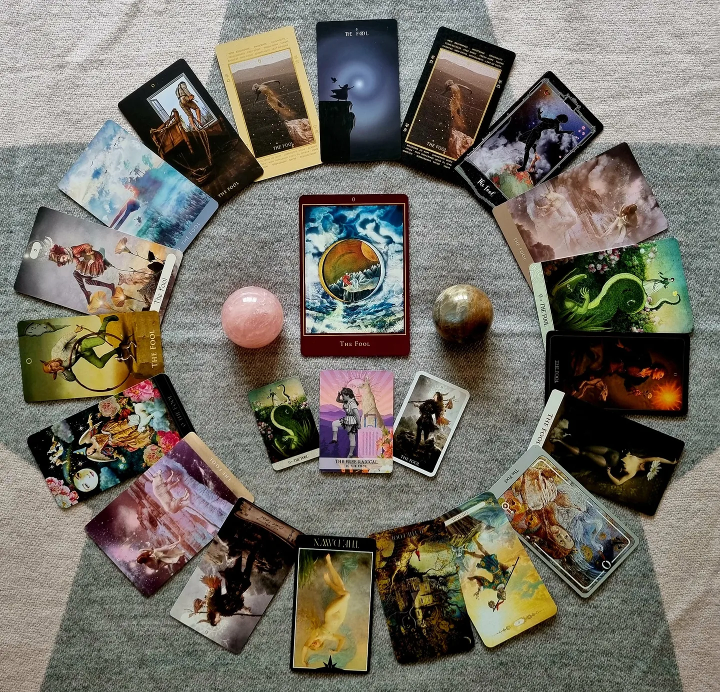 A circle of Fool tarot cards from many decks laid out around a central Fool card with rose quartz and tiger's eye spheres