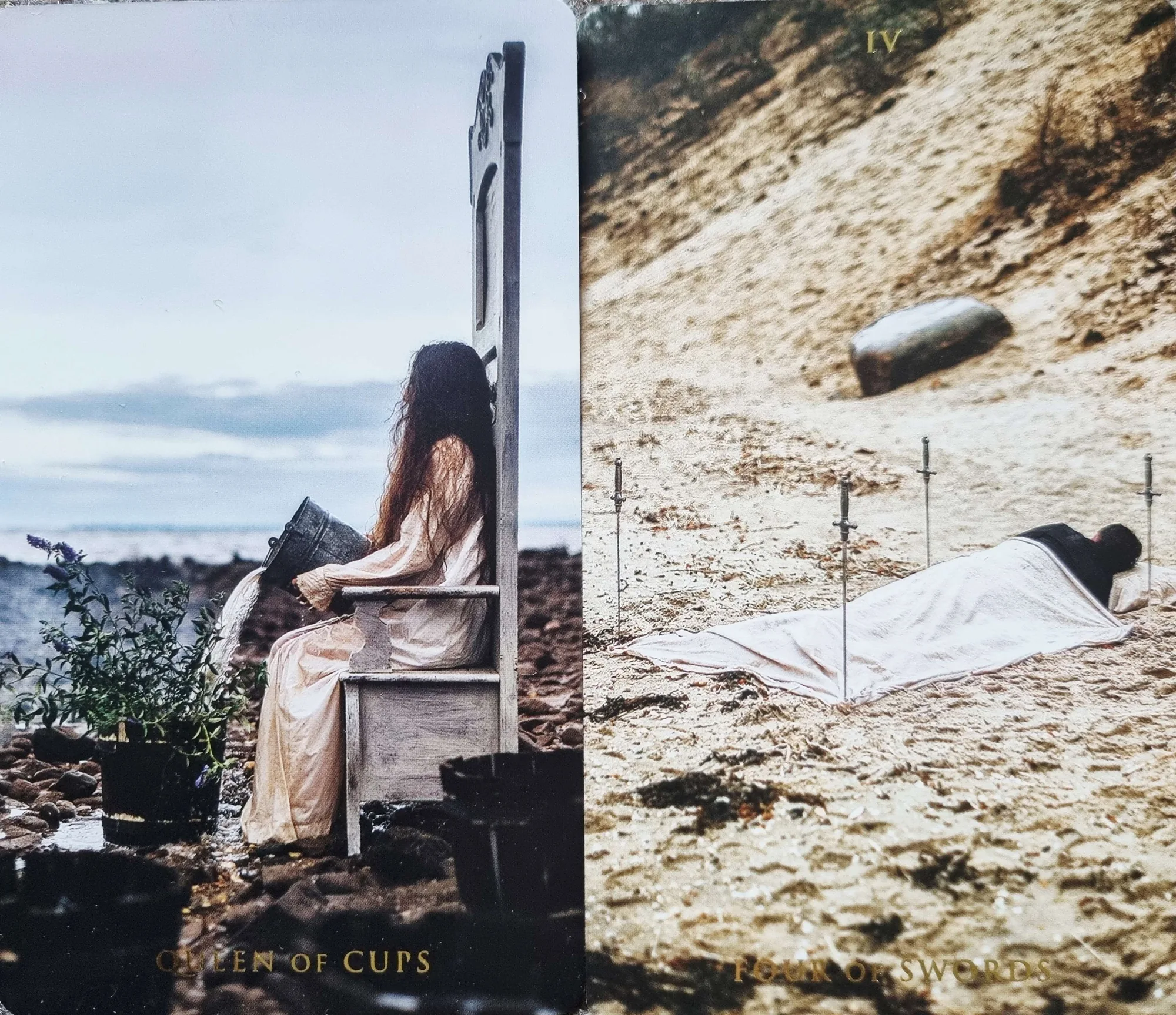 The Queen of Cups and the Four of Swords from the Somnia Tarot side by side, a woman seated on a throne by the sea and a figure lying under a sheet among swords on the sand