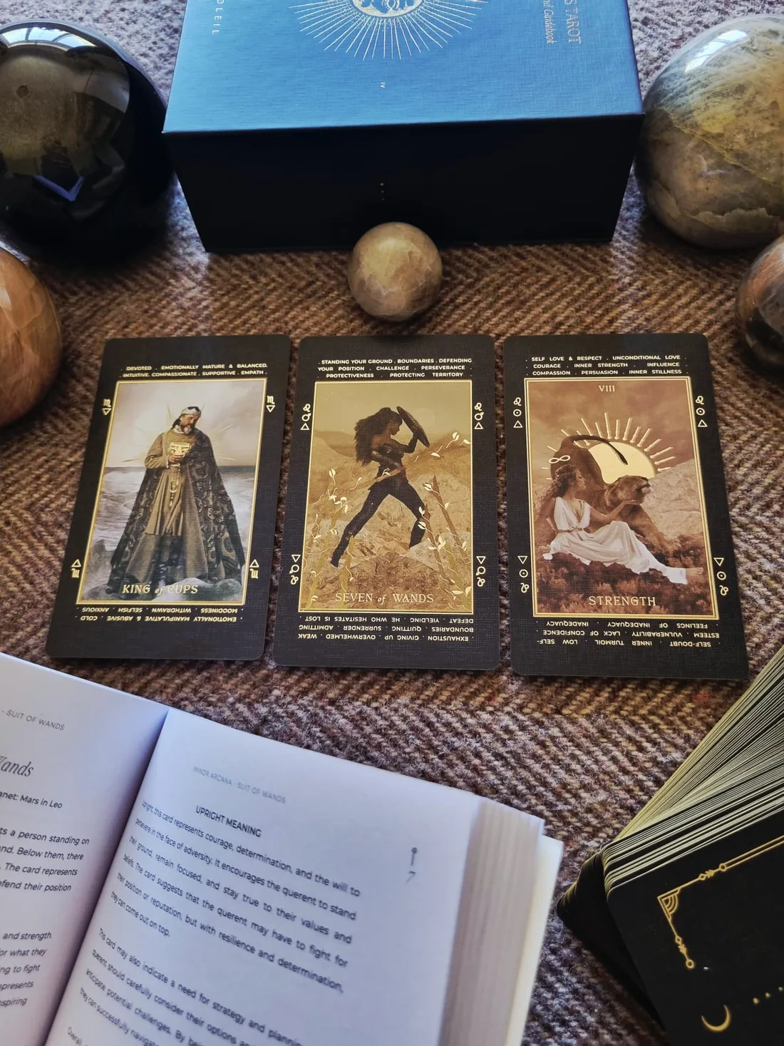 A three card tarot spread beside an open guidebook, the King of Cups, Seven of Wands and Strength