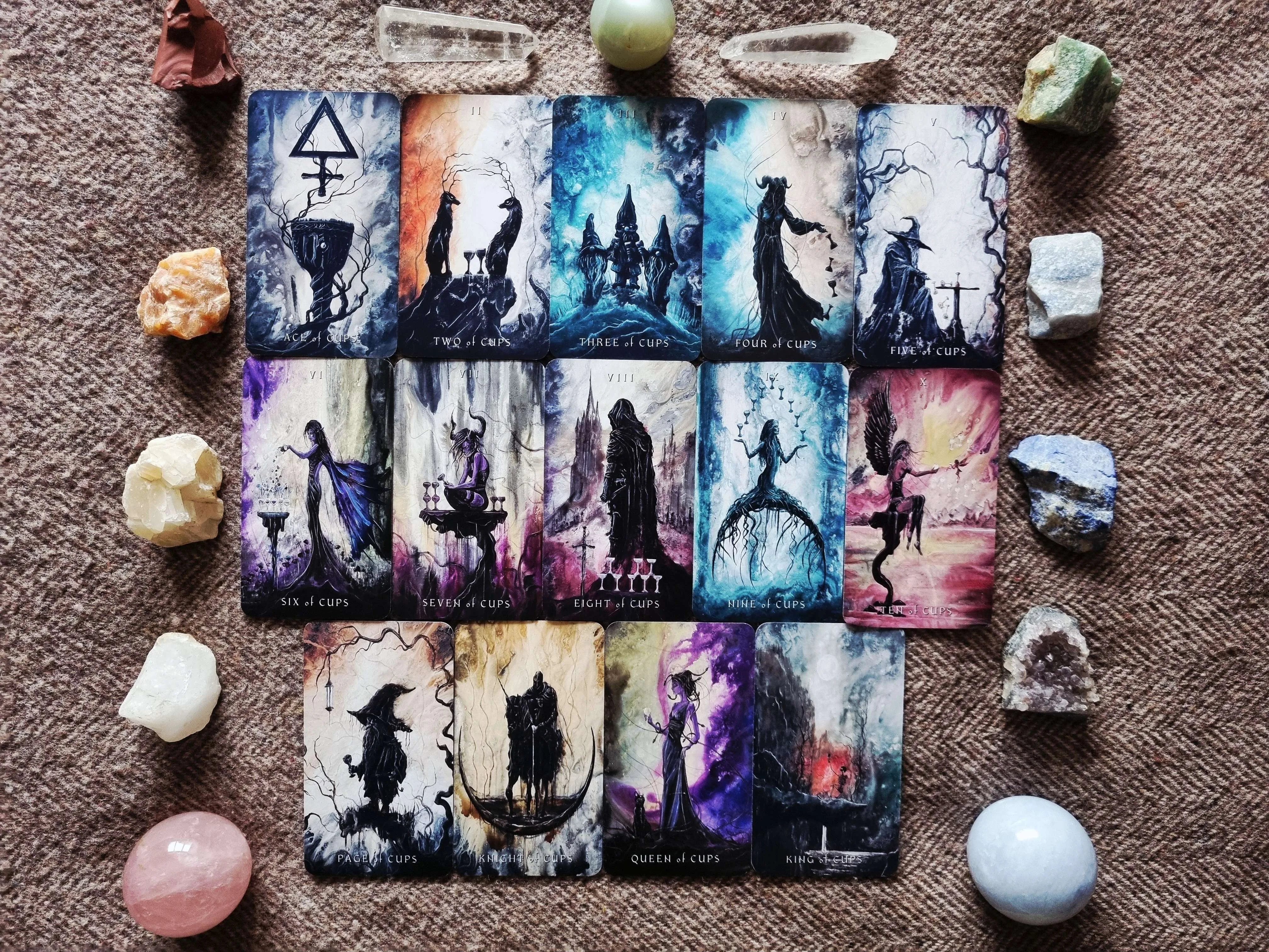 Full Suit of Cups from The Shadows We Cast tarot laid out with crystals