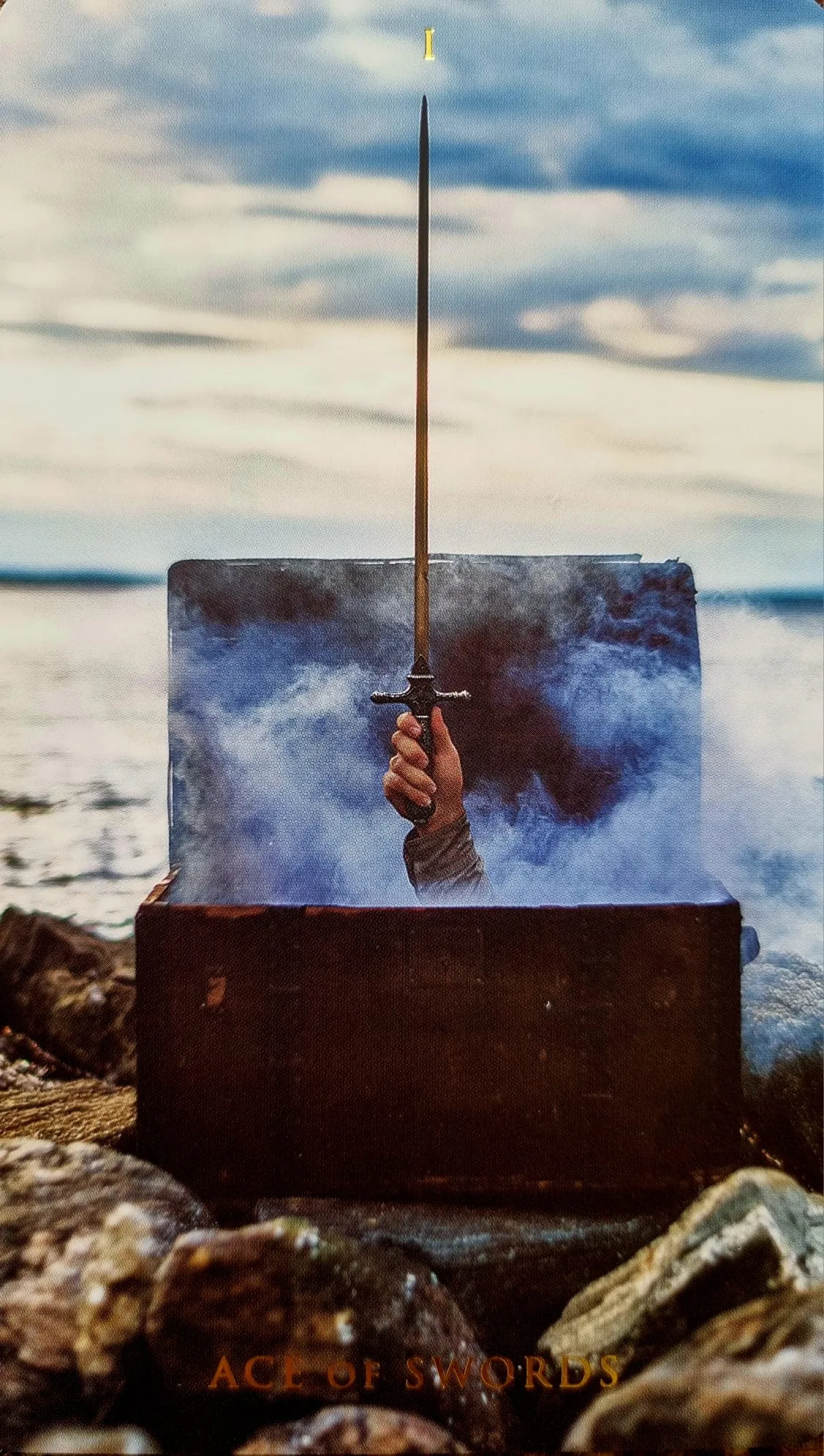 Ace of Swords card: a hand rising from crashing waves holding a sword aloft