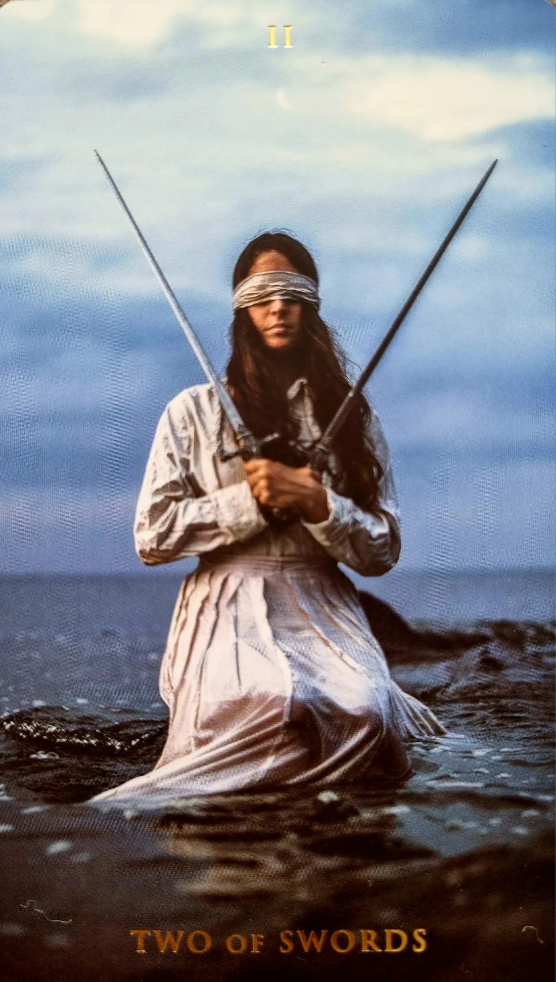 Two of Swords card: a blindfolded figure kneeling in the sea holding two crossed swords