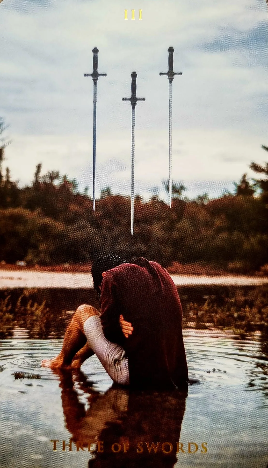 Three of Swords card: three swords hanging above a grieving cloaked figure kneeling in water