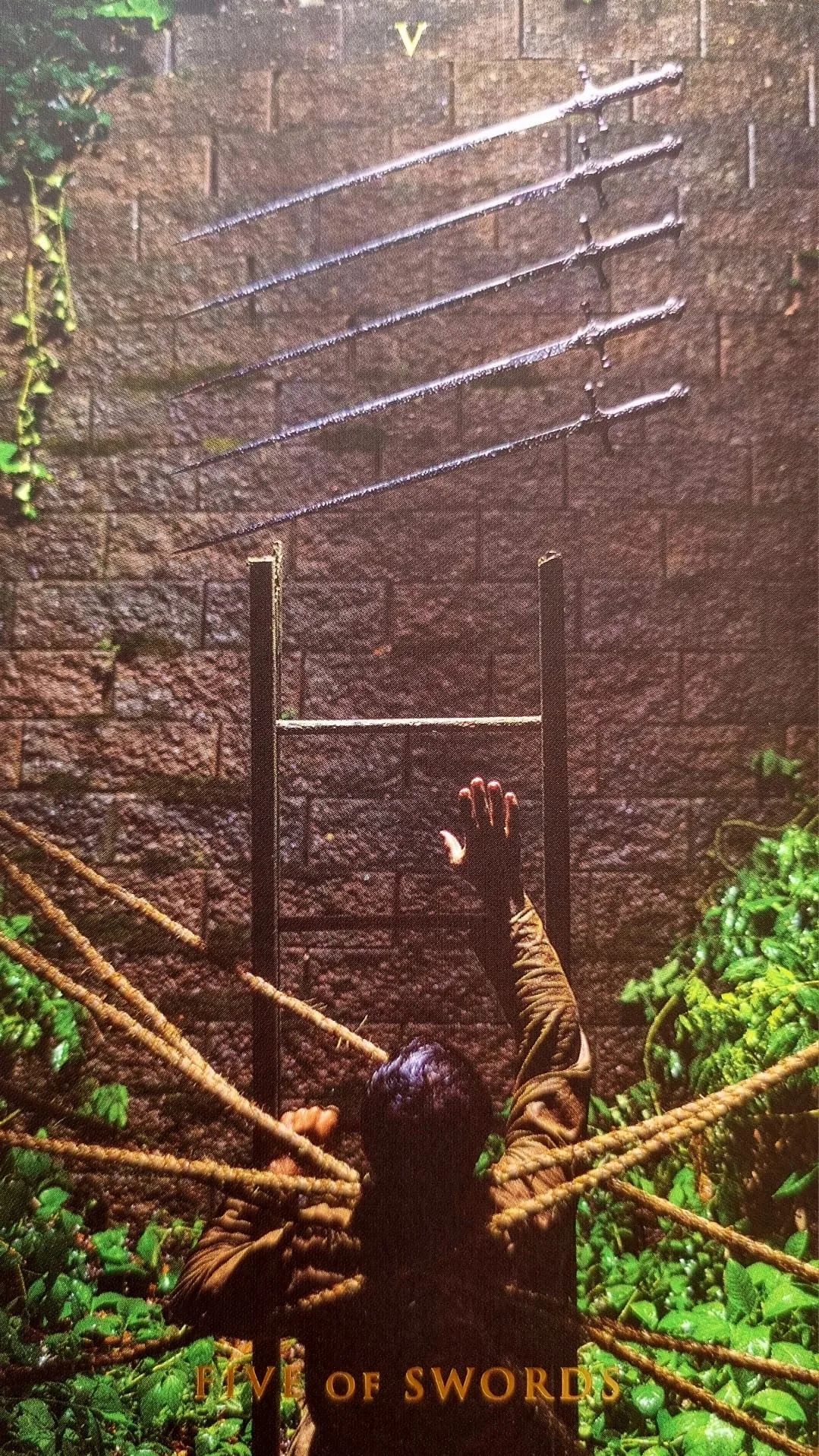 Five of Swords card: a bound figure reaching towards five swords fixed along a rain-soaked wall