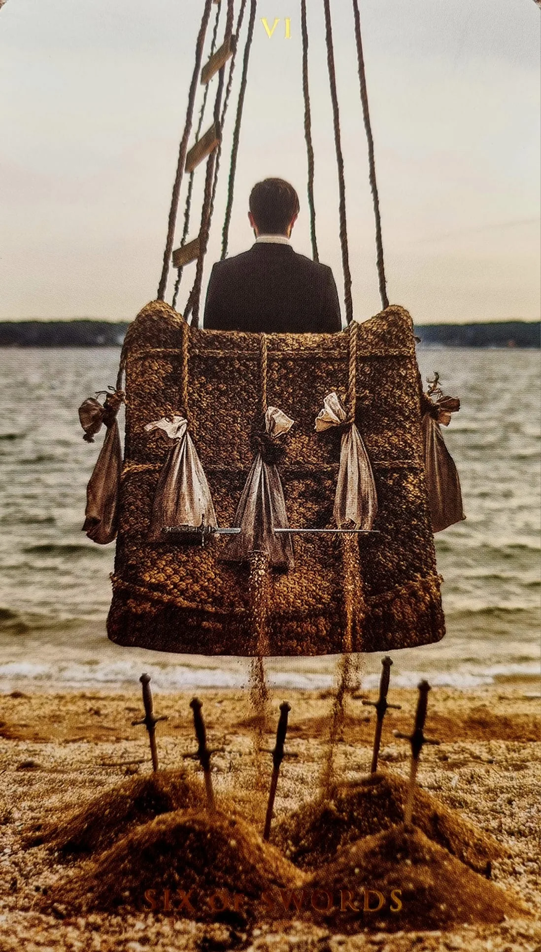 Six of Swords card: a figure being carried away over water in a swaying basket, six swords planted in the sand below