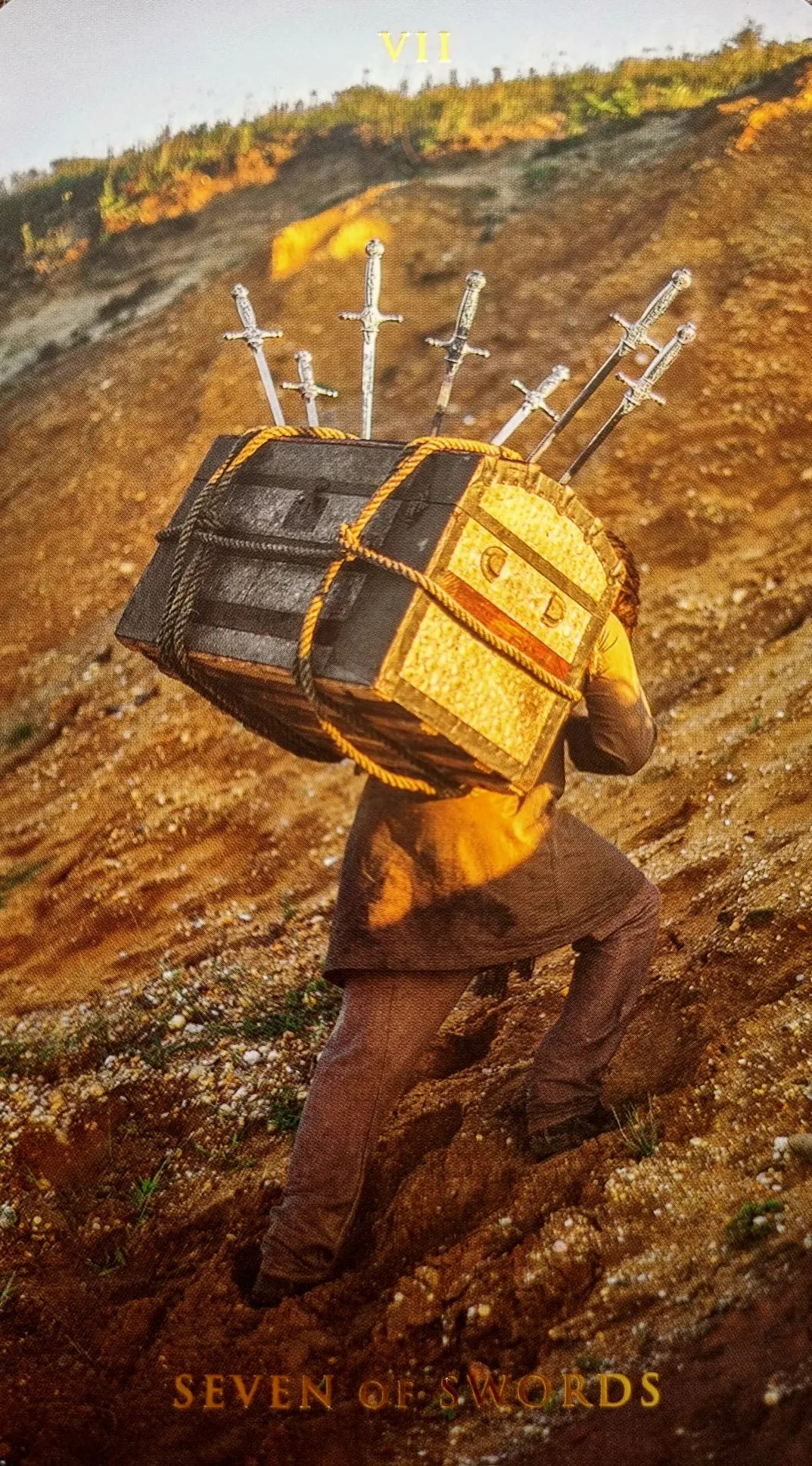 Seven of Swords card: a figure climbing a hillside carrying a chest bristling with swords