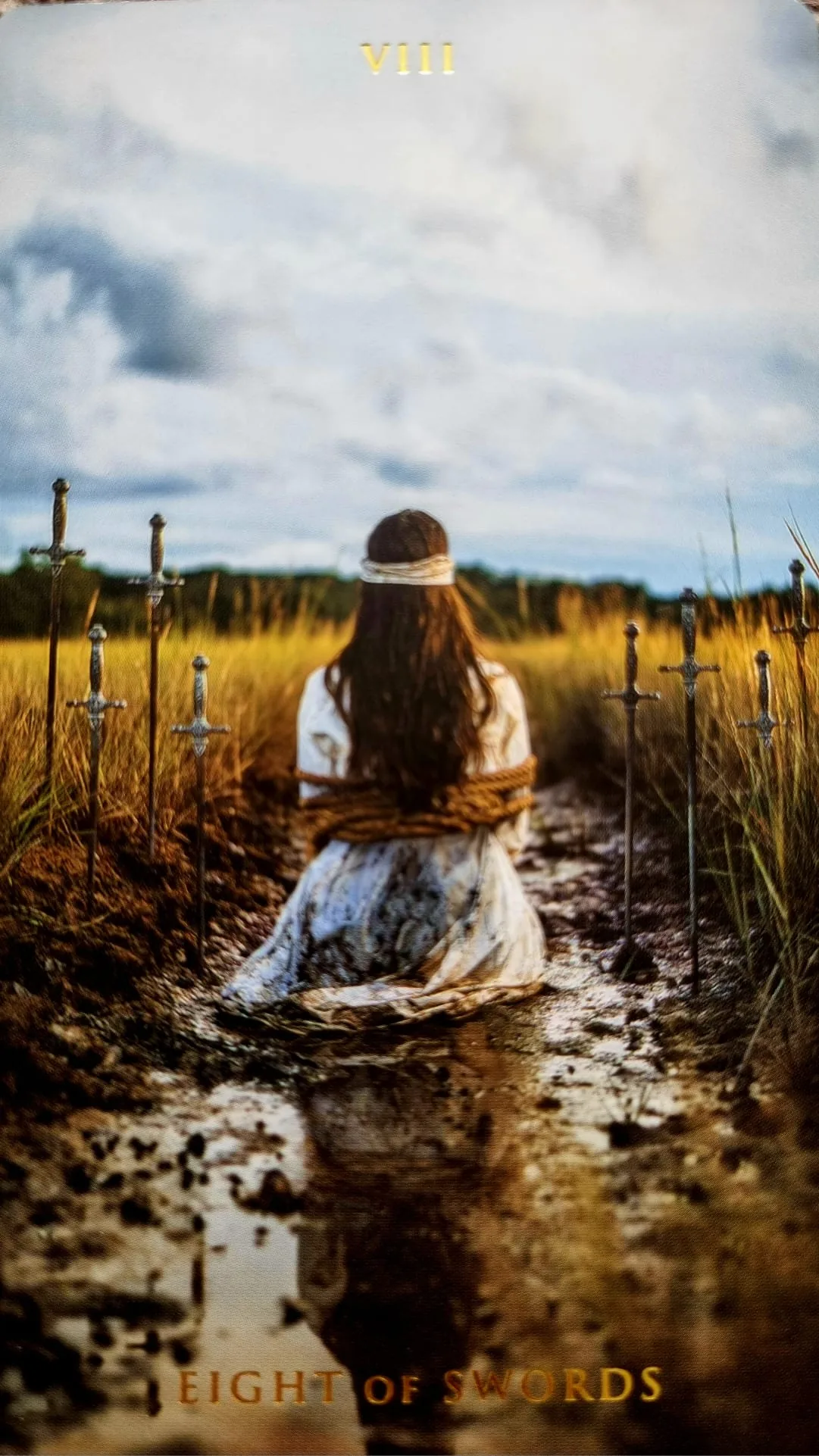 Eight of Swords card: a bound, blindfolded figure kneeling in a muddy path surrounded by eight swords