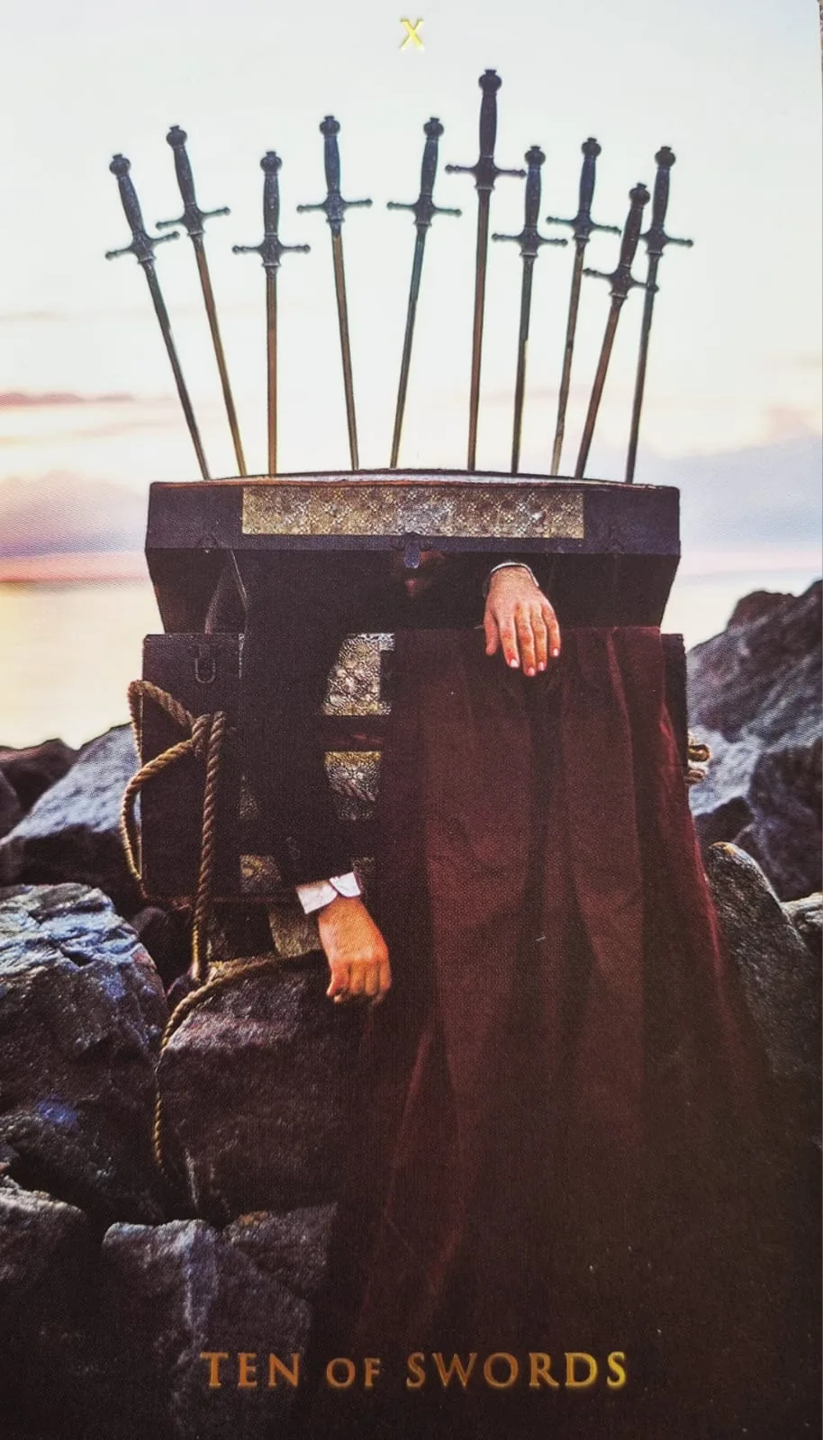 Ten of Swords card: a hooded figure in a red robe pinned beneath ten swords on a rocky shore
