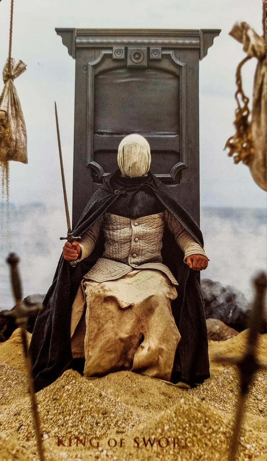 King of Swords card: a robed figure seated on a throne in the sand holding a sword upright