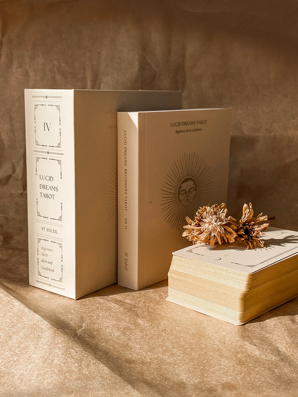 Two boxed editions of the beige Lucid Dreams Tarot by ST SOLEIL stand upright beside a stack of cards and dried flowers on kraft paper
